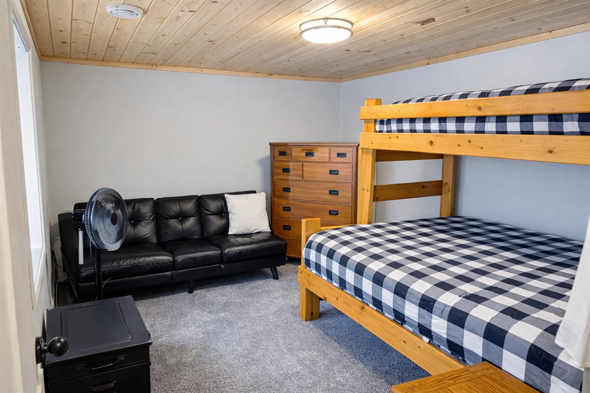 This downstairs bedroom features a full-over-queen bunk bed, a futon couch for lounging, a large dresser for storage, and a fan to keep things cool. It’s a comfy, practical space to relax after a day out, perfect for family or friends.