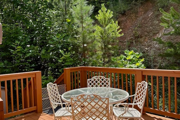 Cozy deck with glass table & white chairs. Lush greenery.