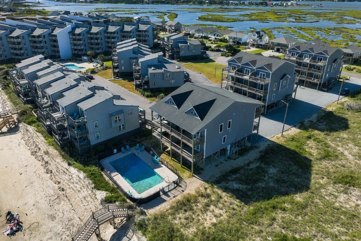 High-angle aerial photograph showcasing the community grounds, featuring an in-ground outdoor pool, sun loungers, private boardwalk beach access over the dunes, and the surrounding coastal landscape.   