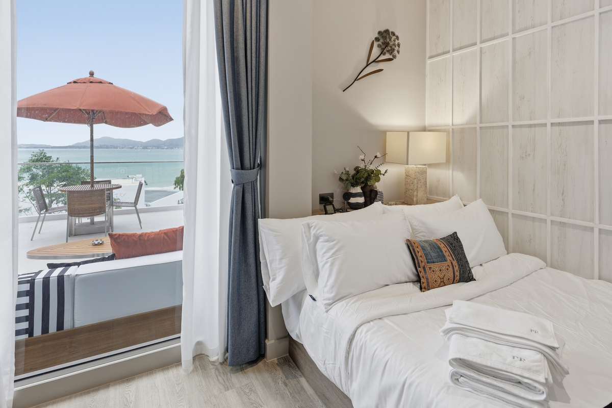 Bedroom with white linens, ocean view balcony, red umbrella, and coastal landscape visible through glass doors.