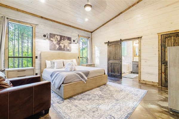 Elegant master bedroom in a luxury cabin, showcasing a spacious bed, high vaulted ceilings, barn doors, and scenic woodland views. Ideal for a peaceful getaway in nature.