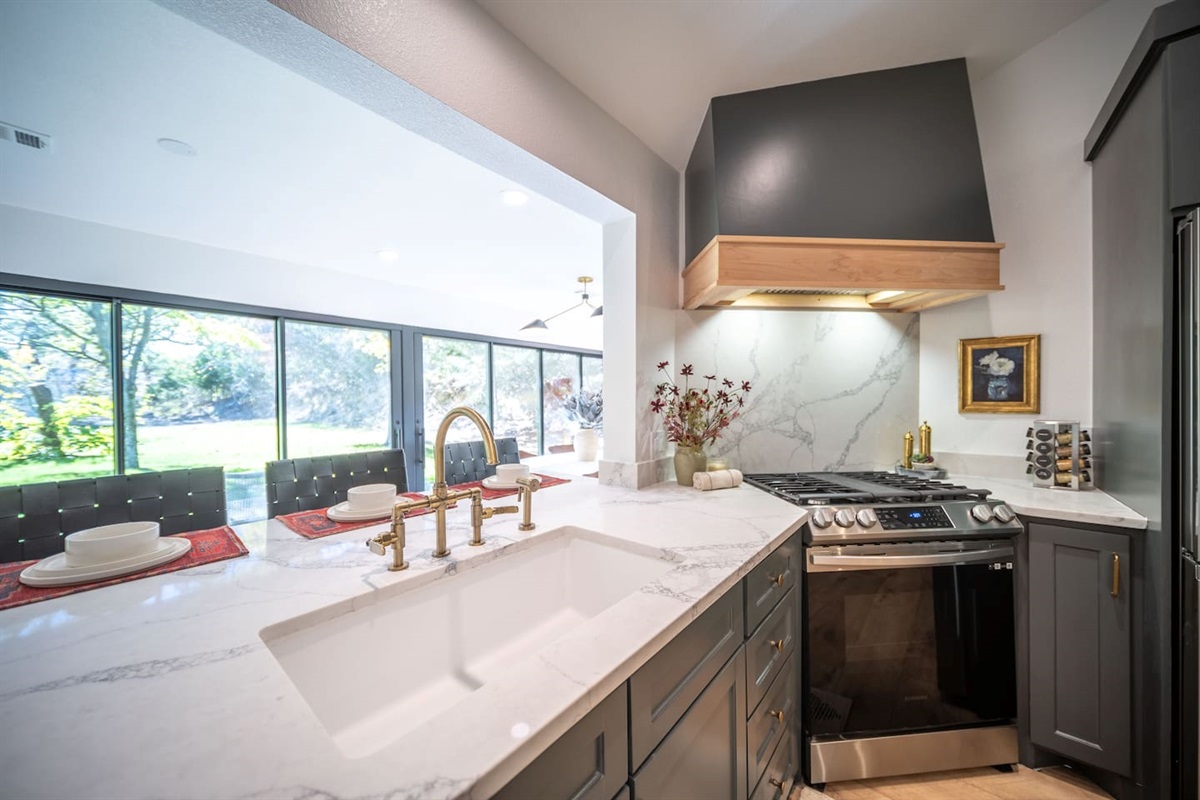 Kitchen perspective featuring the gas range, custom hood, marble backsplash, and island sink with breakfast bar seating and a wall of glass for a wash of natural light throughout. The dining room is just on the other side. Fully stocked kitchen!