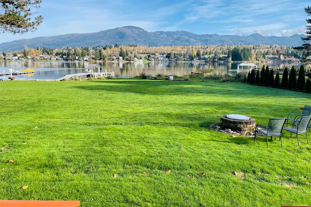 A wide, green backyard stretches to the lake, with a dock and fire pit ready for evenings outdoors. The open lawn feels inviting, and the calm water reflects the shoreline, creating a peaceful connection between land and lake.