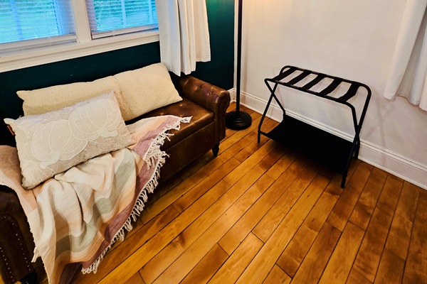 Cozy layers and garden views—our cushioned bench in Queen’s Landing invites you to curl up and unwind. Soft blankets, plush pillows, a handy luggage rack, and those gorgeous hardwood floors complete the scene.