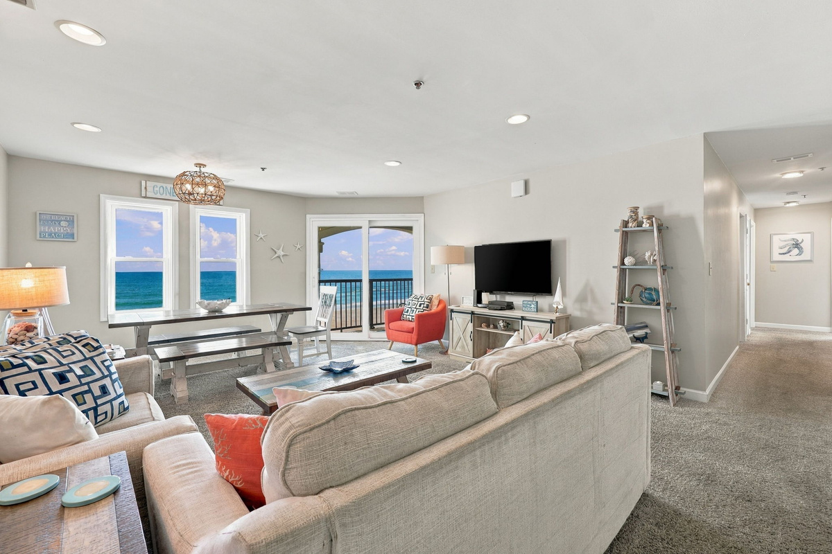 Expansive view of the main living area showing plush sofa seating, a coastal dining table with bench seating, a ladder display shelf, and dual window cutouts alongside sliding glass doors revealing stunning Atlantic views.   