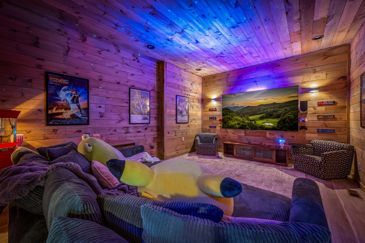 Cozy movie room with a big screen, plush seating, video game consoles, a popcorn machine, and LED lighting for an immersive movie night experience.