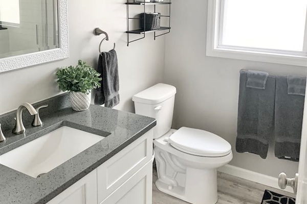 Refresh in this bright, updated bathroom featuring a sleek grey quartz vanity top and a white shaker-style cabinet. The space is finished with clean white walls, dark charcoal towels, and a window that provides plenty of natural light.