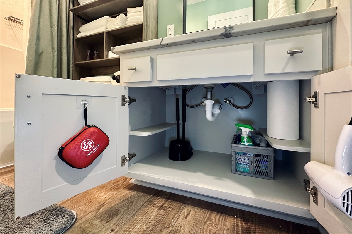Hall bathroom under-sink cabinet stocked with cleaning supplies, paper towels, a first aid kit, and hair dryer for added guest convenience.
