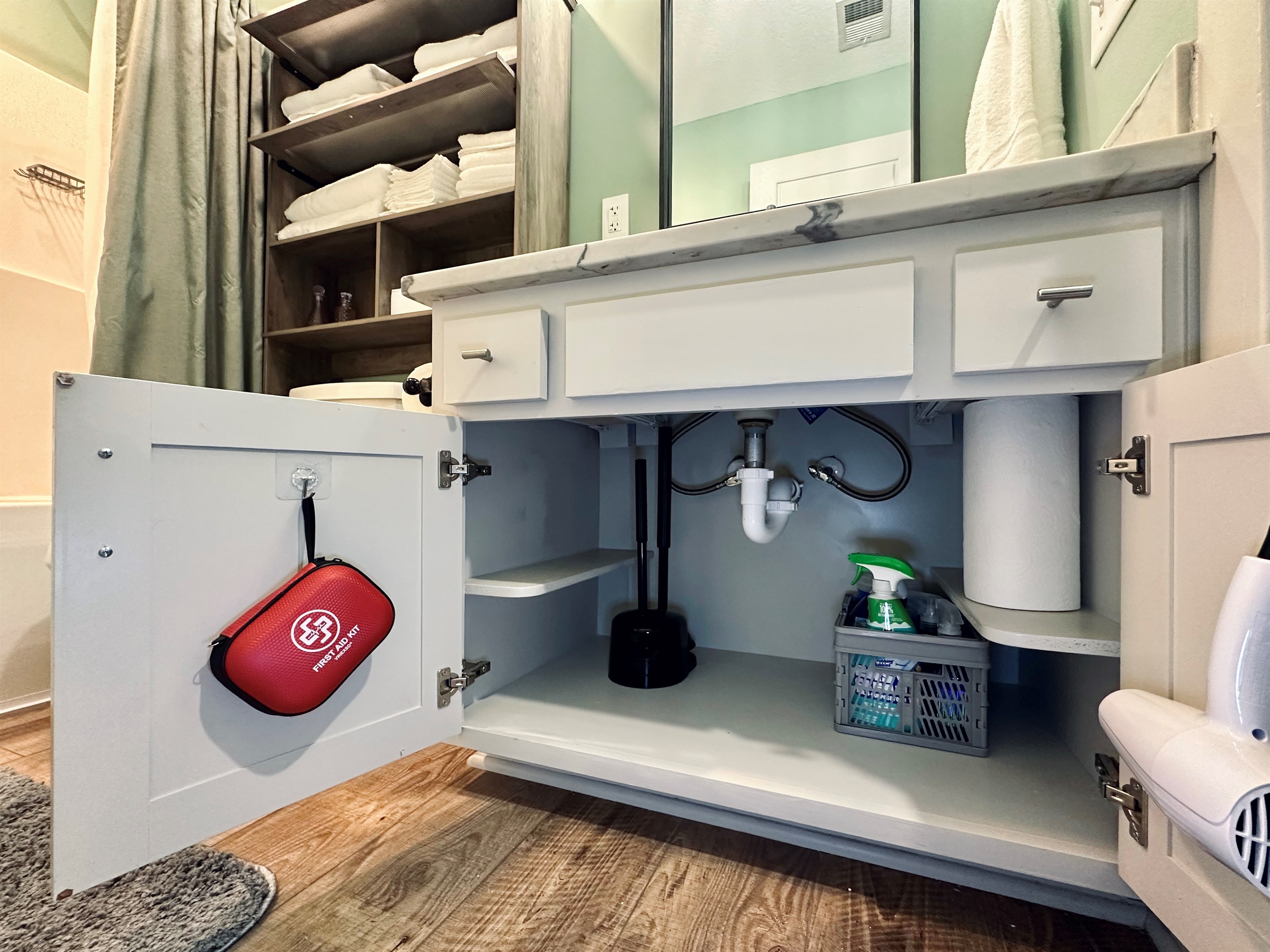 Hall bathroom under-sink cabinet stocked with cleaning supplies, paper towels, a first aid kit, and hair dryer for added guest convenience.
