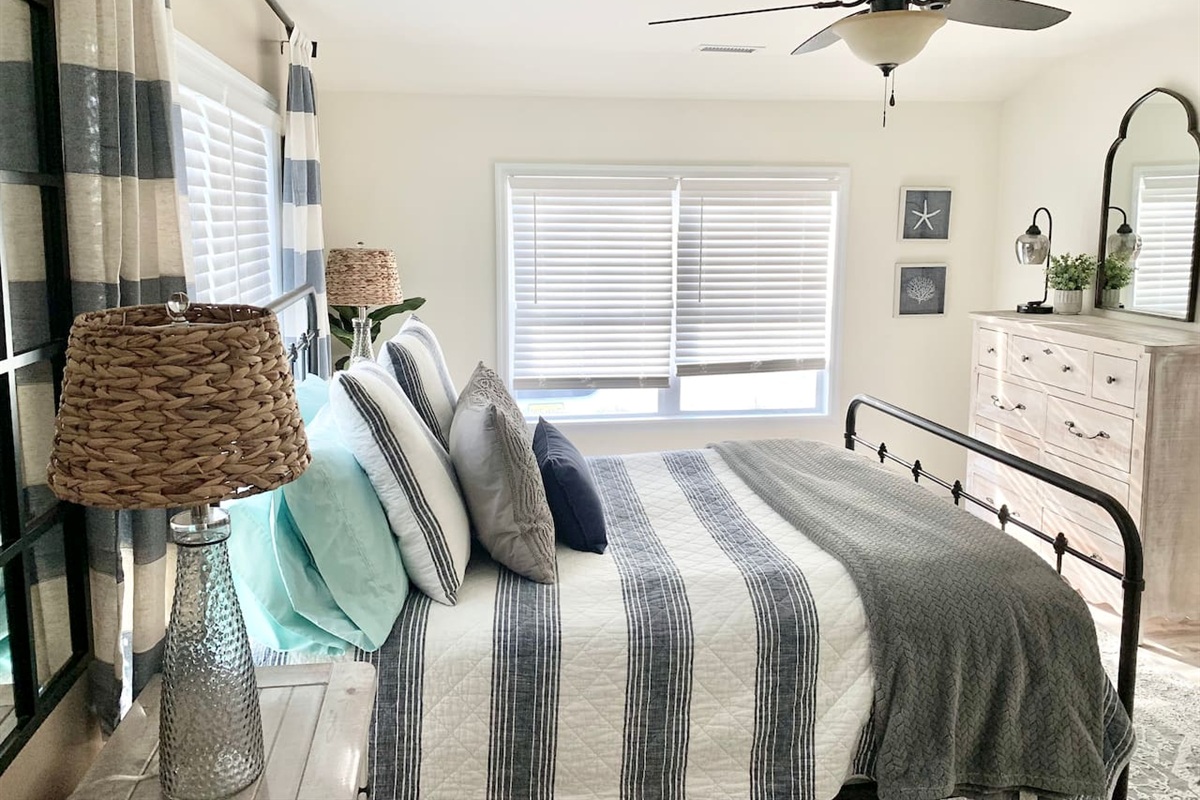 Wake up in a sun-drenched sanctuary featuring a black iron bed frame and plush nautical-themed bedding. This bright primary bedroom is complete with rustic wood furniture, woven textures, and soft coastal accents for a truly peaceful stay.