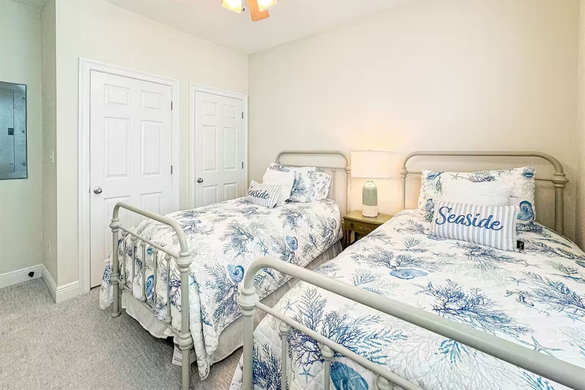 Bright guest bedroom with two twin beds, coastal-themed linens, and plenty of natural light. Includes a ceiling fan and cozy setup—perfect for kids or friends.