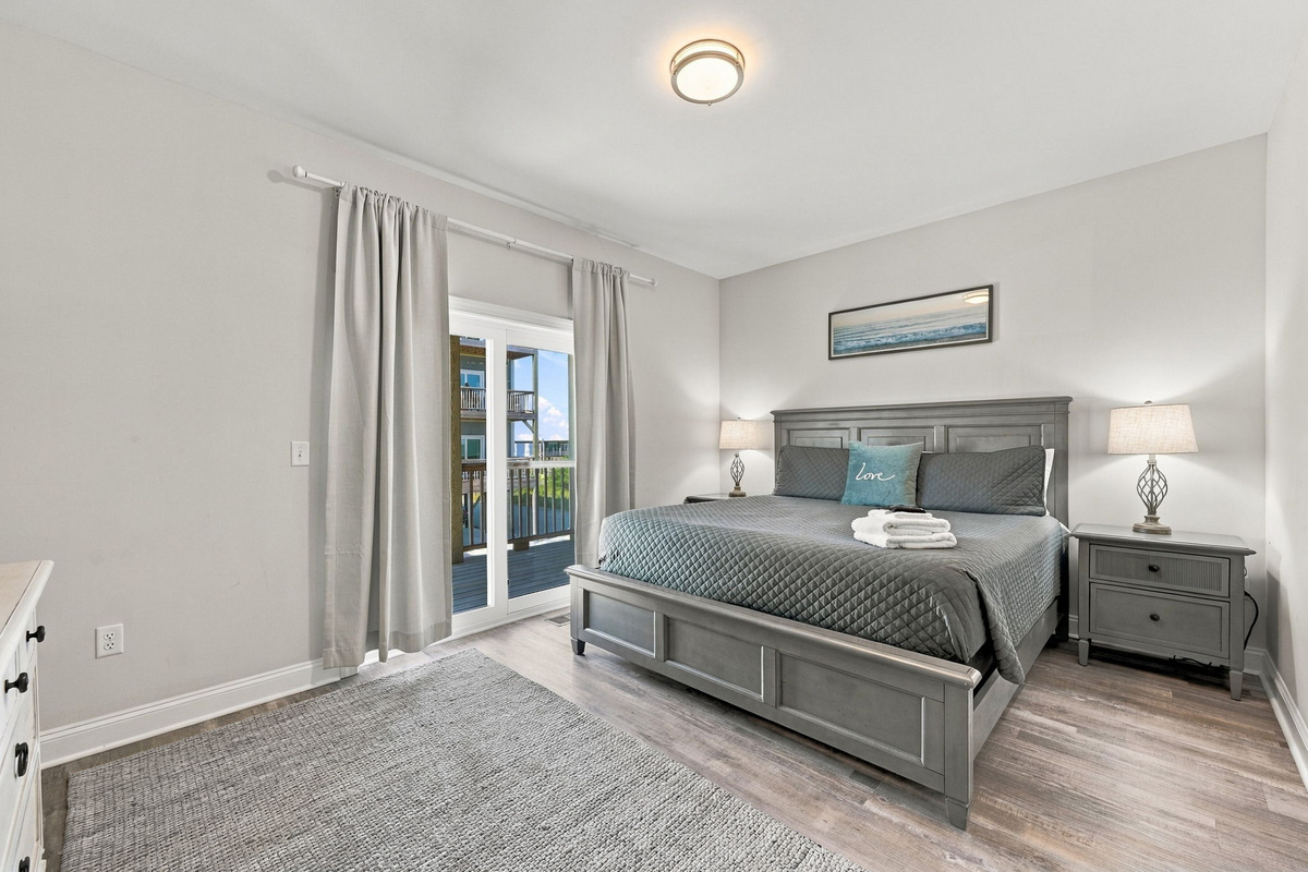 Primary master bedroom featuring a dark grey wooden king bed set, grey quilted bedding with coastal accent pillows, matching nightstands, and sliding glass doors leading out to a private balcony.   
