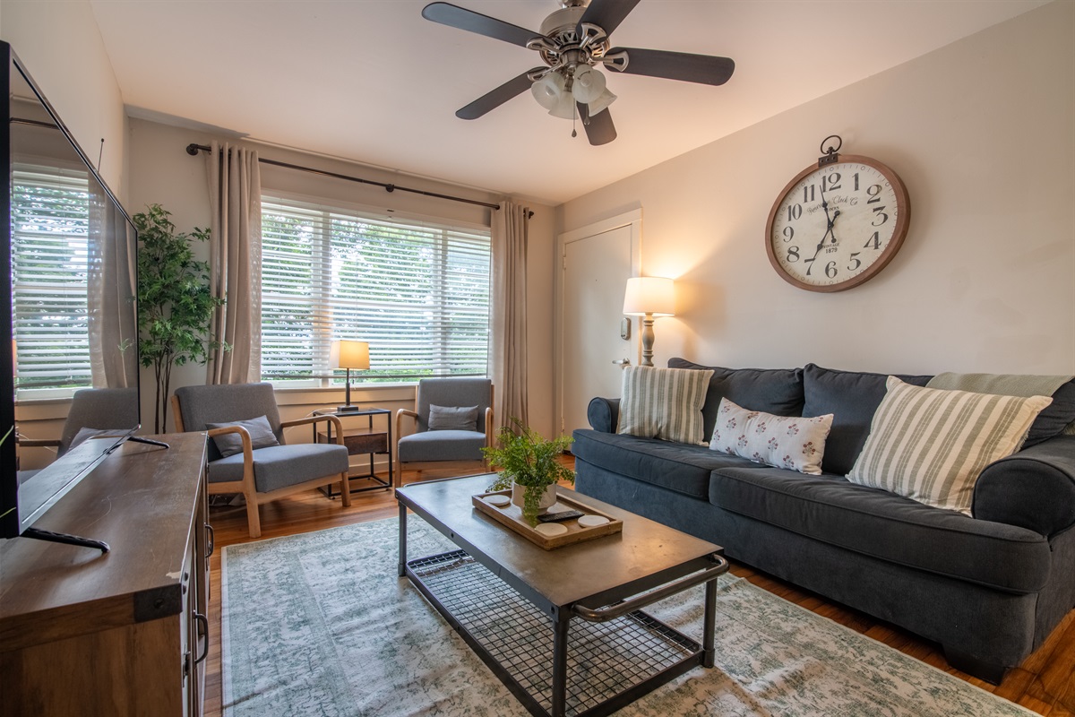 Cozy, sun-filled living room with large windows, ceiling fan, and stylish decor—perfect for gathering or relaxing.