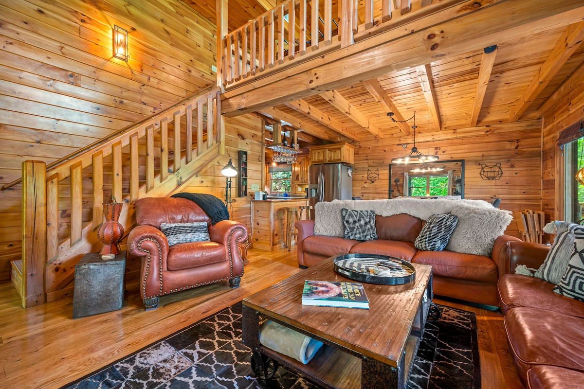 Warm and inviting main living room with rich wood interiors, cozy leather seating, and an open-concept layout connecting seamlessly to the kitchen and loft above. 