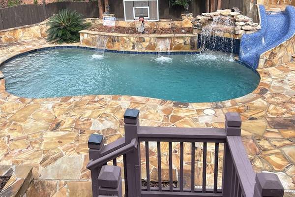 Resort-style backyard pool with natural stone decking, rock waterfall, built-in slide, and basketball hoop. Surrounded by mature trees for privacy, perfect for relaxing, entertaining, or family fun in a peaceful outdoor setting.