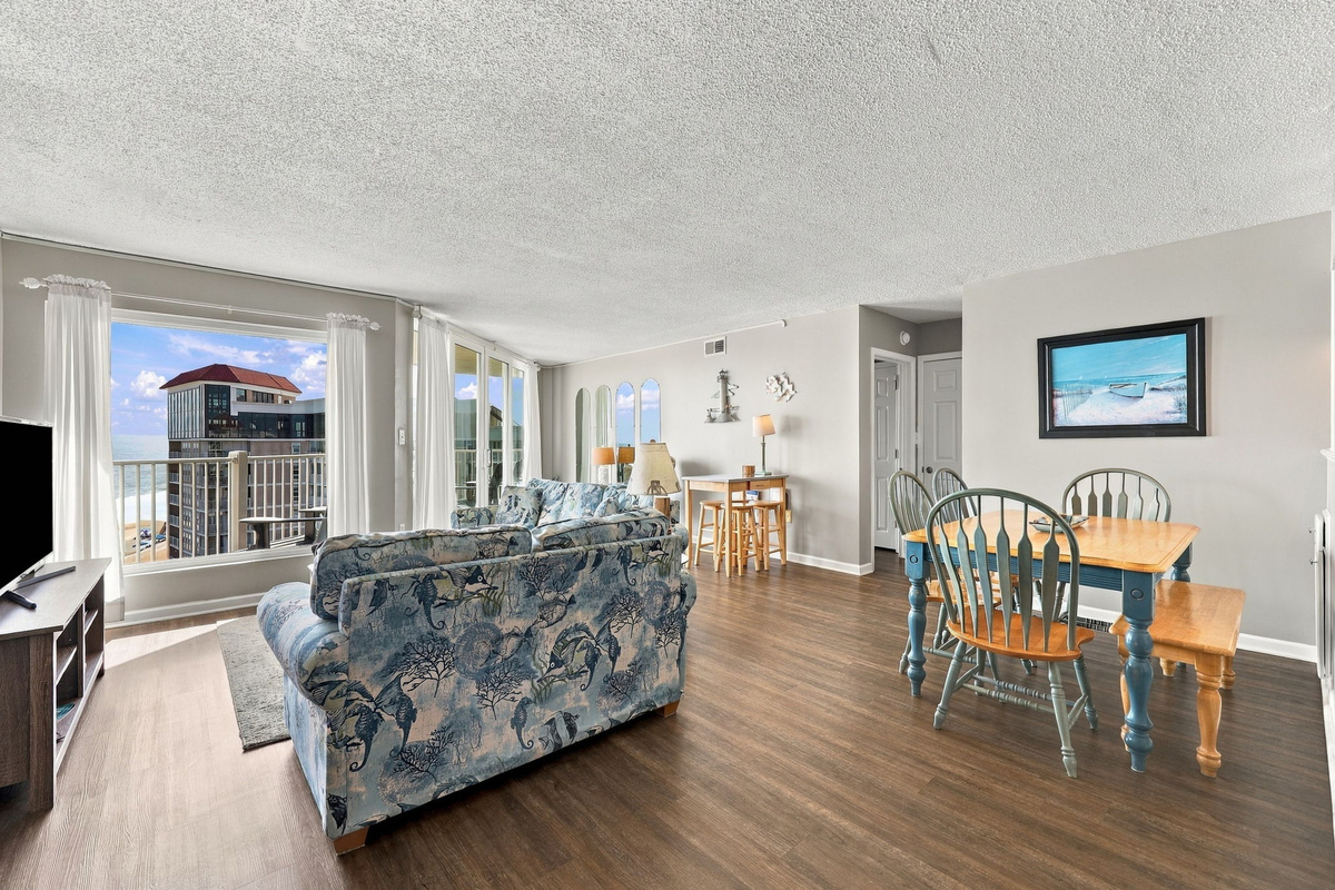 Spacious living area featuring a floral patterned sofa, wooden dining table with chairs, and sliding glass doors leading to the balcony with ocean views.