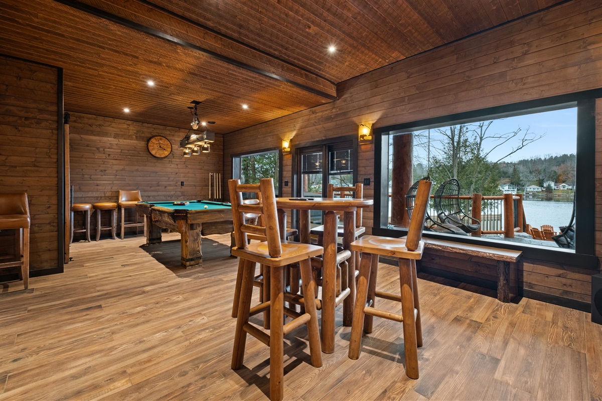 Spacious lower-level games room featuring rustic log bar-height seating, pool table, and large picture window overlooking the lake.