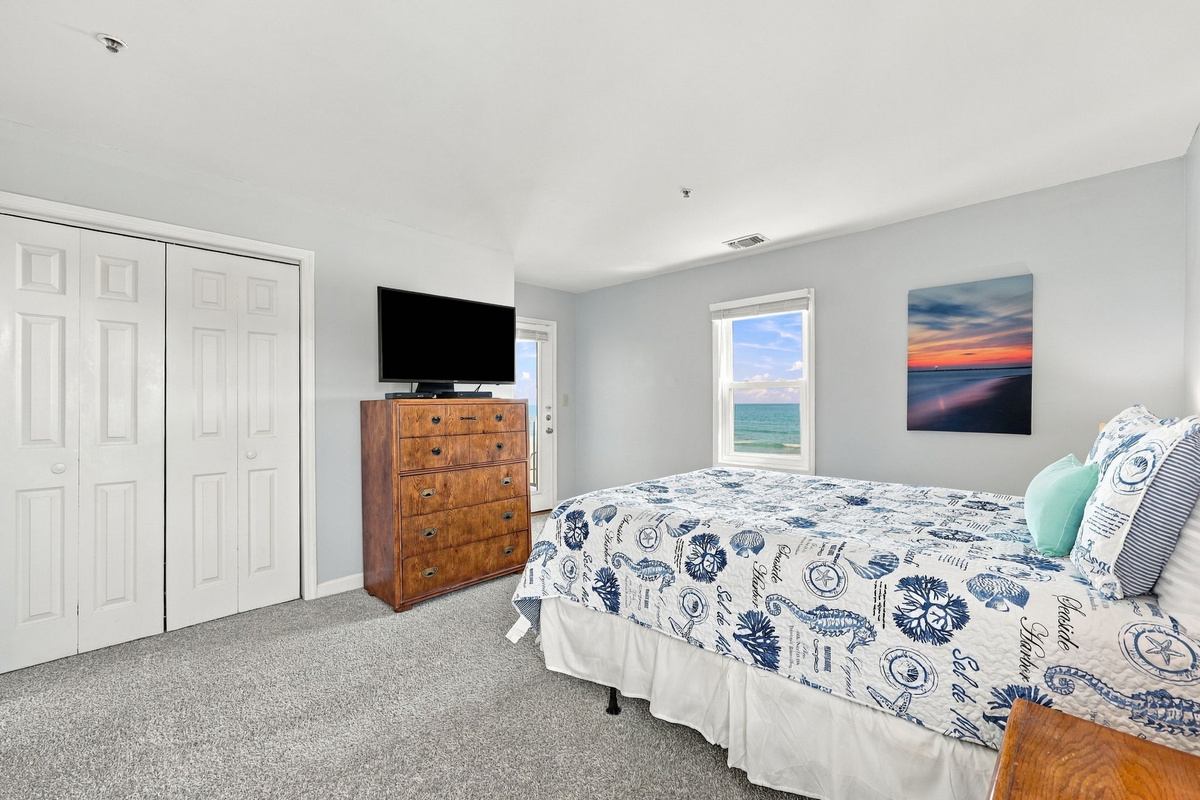 Guest bedroom view showing a cozy bed in blue-and-white sea life linens, a tall warm-wood dresser with TV, ample bi-fold closet space, vibrant beach sunset art, and a large window framing ocean waters.   