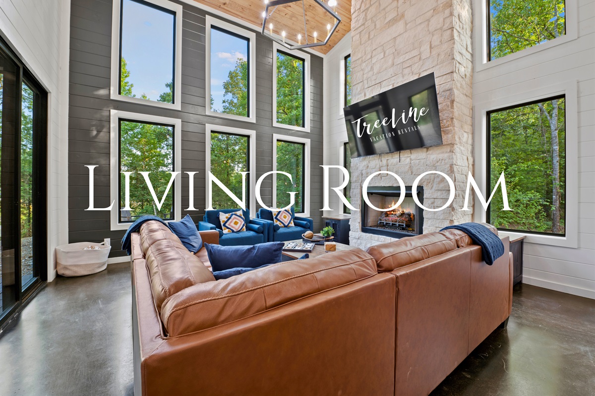Luxuriate in the expansive living area, featuring high ceilings, towering windows, and a comfortable leather sofa centered around a warm fireplace.
