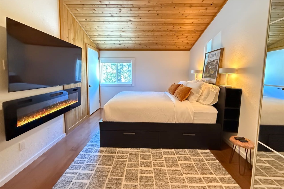 Primary Loft Bedroom: A spacious loft bedroom featuring a comfortable queen-size bed, a modern built-in electric fireplace, a 65" Smart TV, closet with mirrored doors and integrated under-bed storage solutions for your gear.