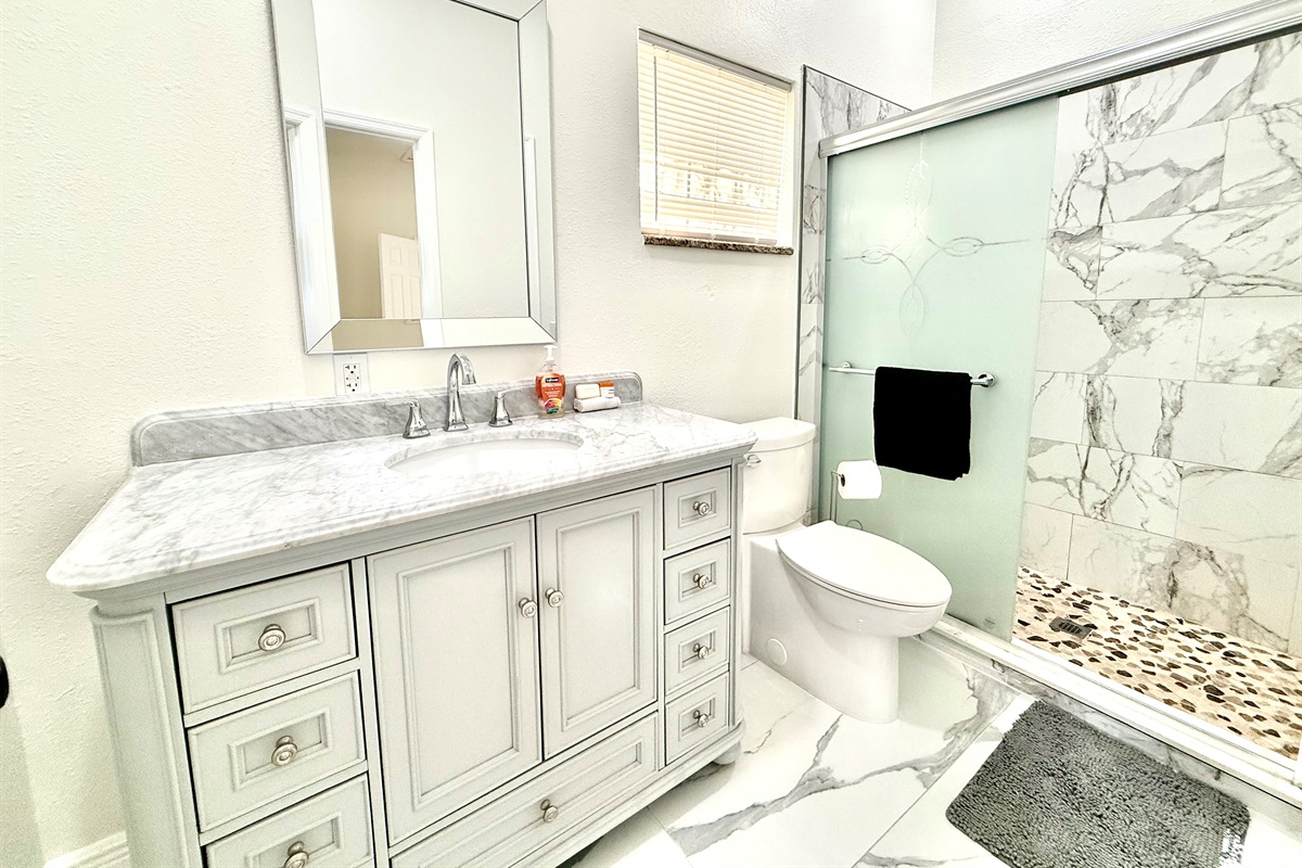 Modern elegance meets function in this beautifully updated bathroom, featuring a sleek marble vanity, stylish gray cabinetry, and a spa-inspired walk-in shower with frosted glass and pebble stone flooring.