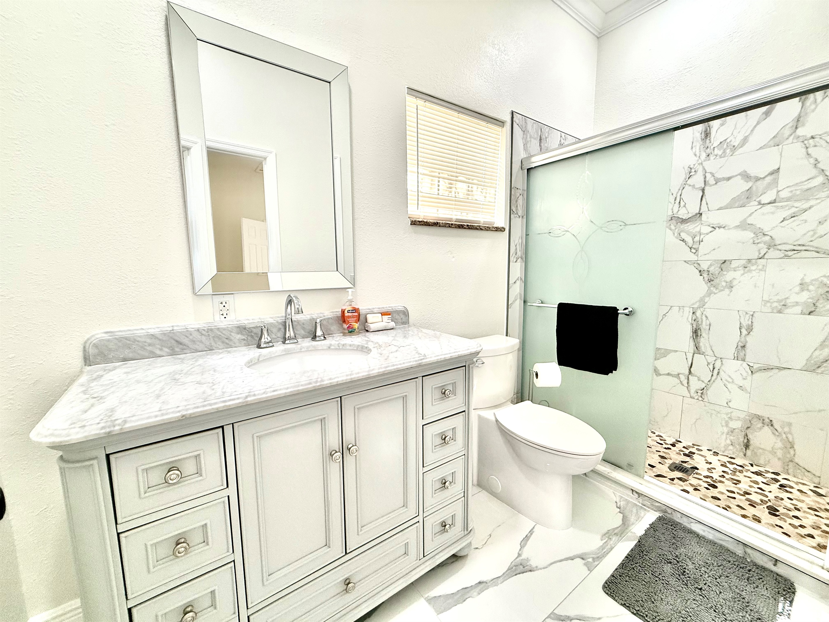 Modern elegance meets function in this beautifully updated bathroom, featuring a sleek marble vanity, stylish gray cabinetry, and a spa-inspired walk-in shower with frosted glass and pebble stone flooring.