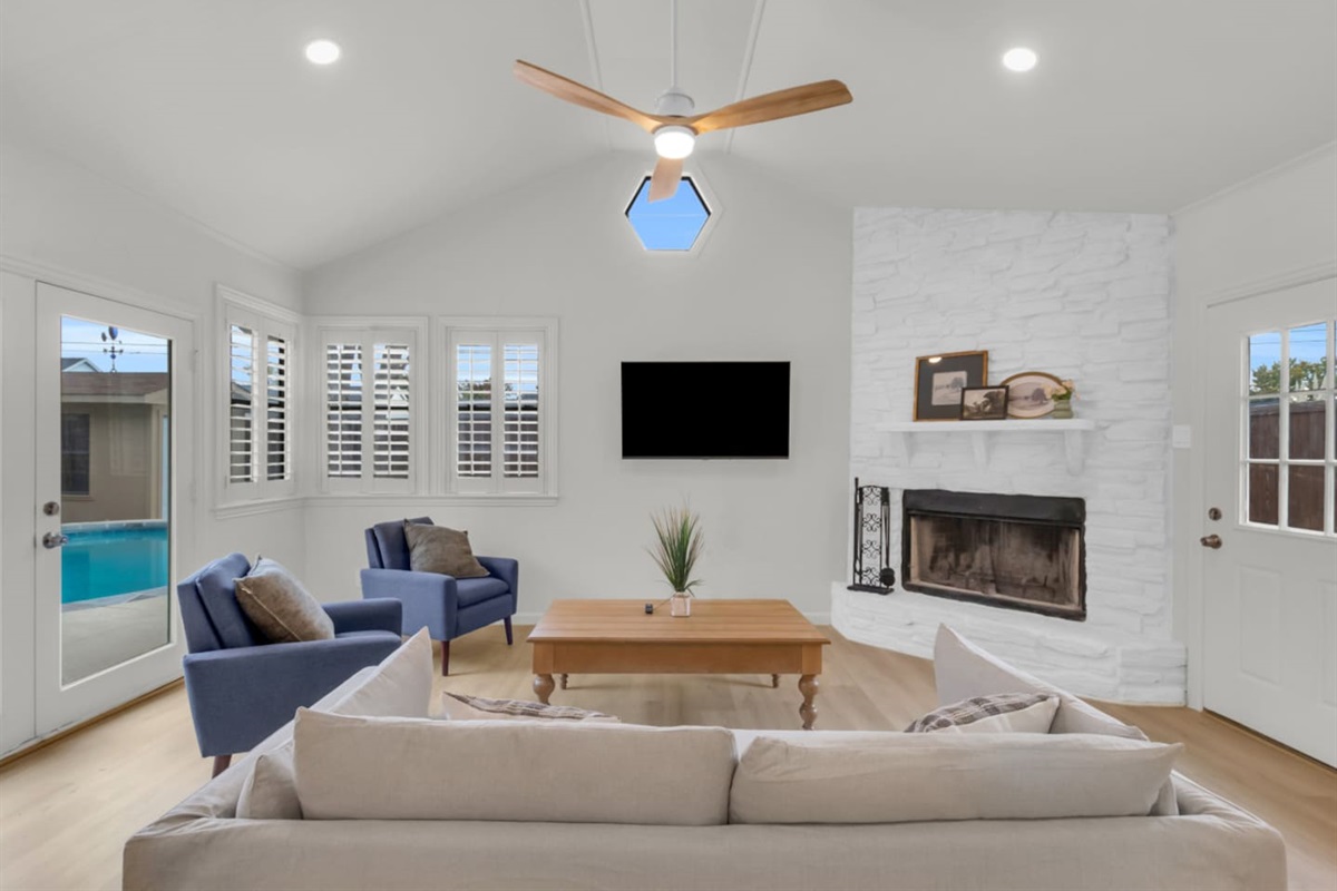 Bright private living room with mounted TV, modern ceiling fan, and a charming indoor fireplace. Direct access to the pool through the private patio doors.