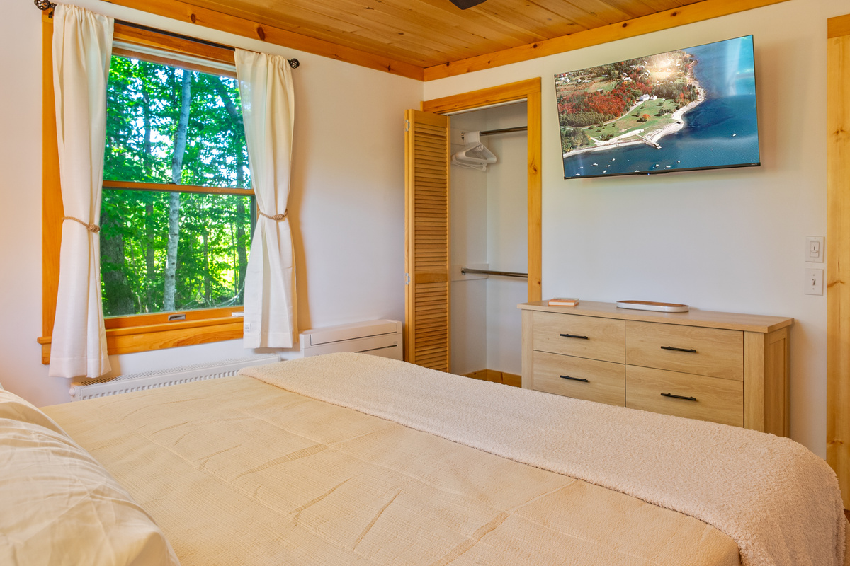 Enjoy the fully equipped primary bedroom, complete with a wall-mounted flat-screen TV and matching light-wood dresser. This view highlights the tongue-and-groove pine ceiling, an open bifold closet, and a large window framing serene forest views.