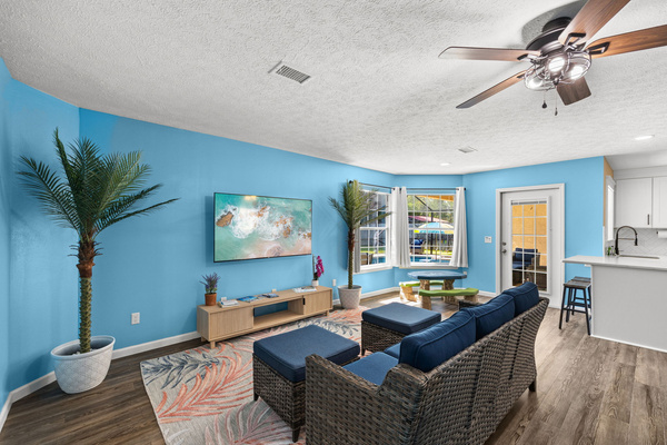A vibrant coastal living room with blue accents, tropical plants, and a 65‑inch TV overlooking the pool area.