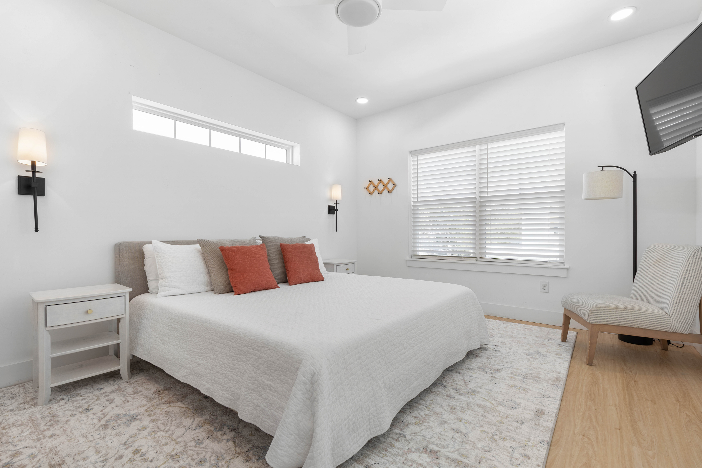 A king bed with sconces to either side, a chair in the corner, and a television on the wall. Rust-colored pillows pick up the warmth in a room otherwise given over to white linen and quiet morning light.