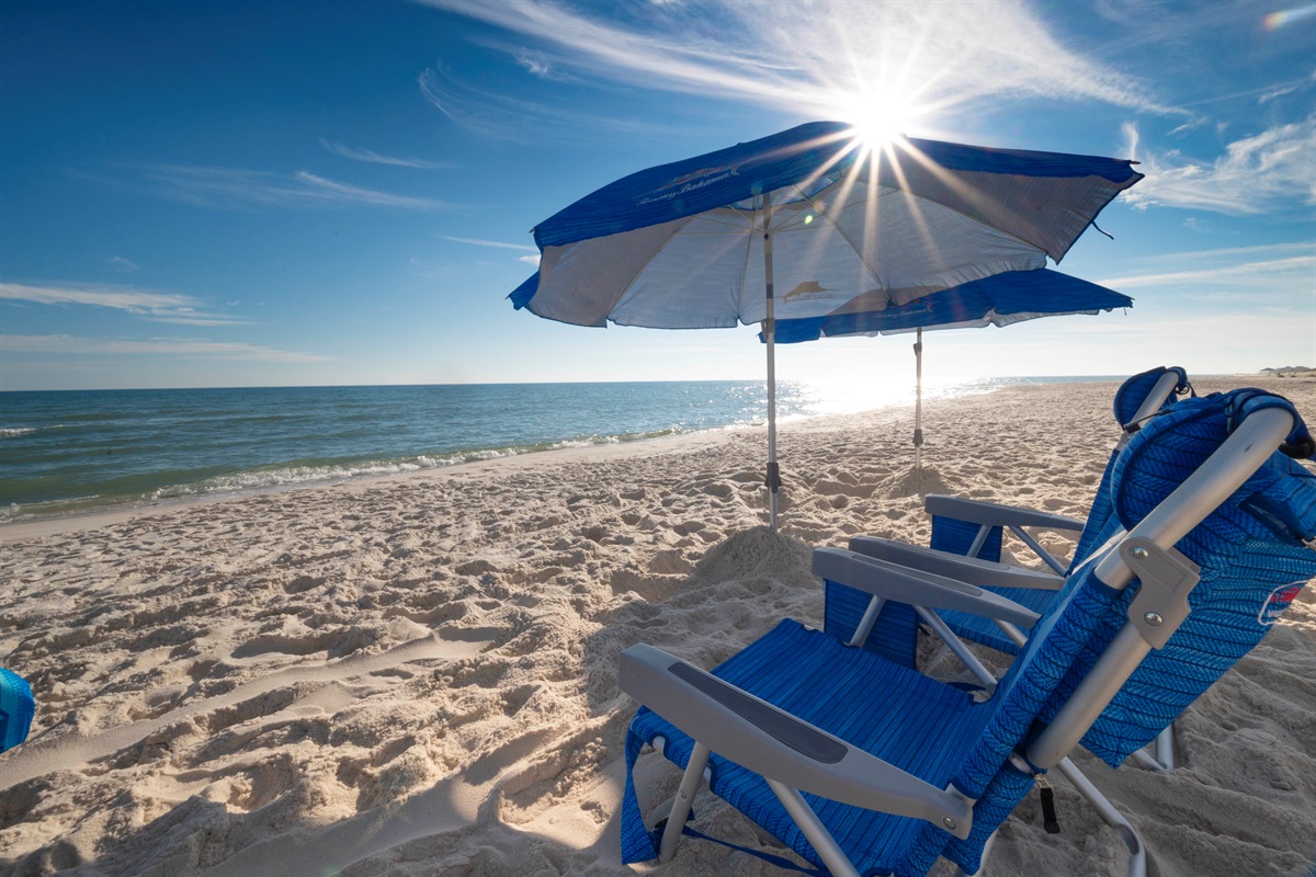 Relax in comfort with blue chairs and an umbrella, basking under sunny skies by calm waves on a sandy shore.