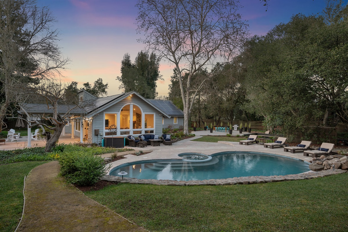 To the left, a pool table with seating outside the living room doors. In front, the pool, rock waterfall, pool loungers, and couches. To the right, the dining pavilion and bocce court.