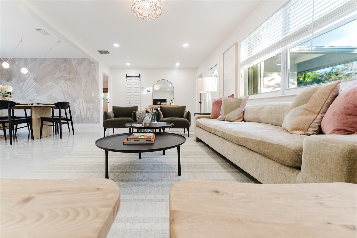 Light-filled living room with a 65" smart TV, sound bar, and subwoofer, featuring plush designer seating, neighborhood views, curated games, and reading materials—perfect for relaxing, entertaining, and family movie nights in West Palm Beach.