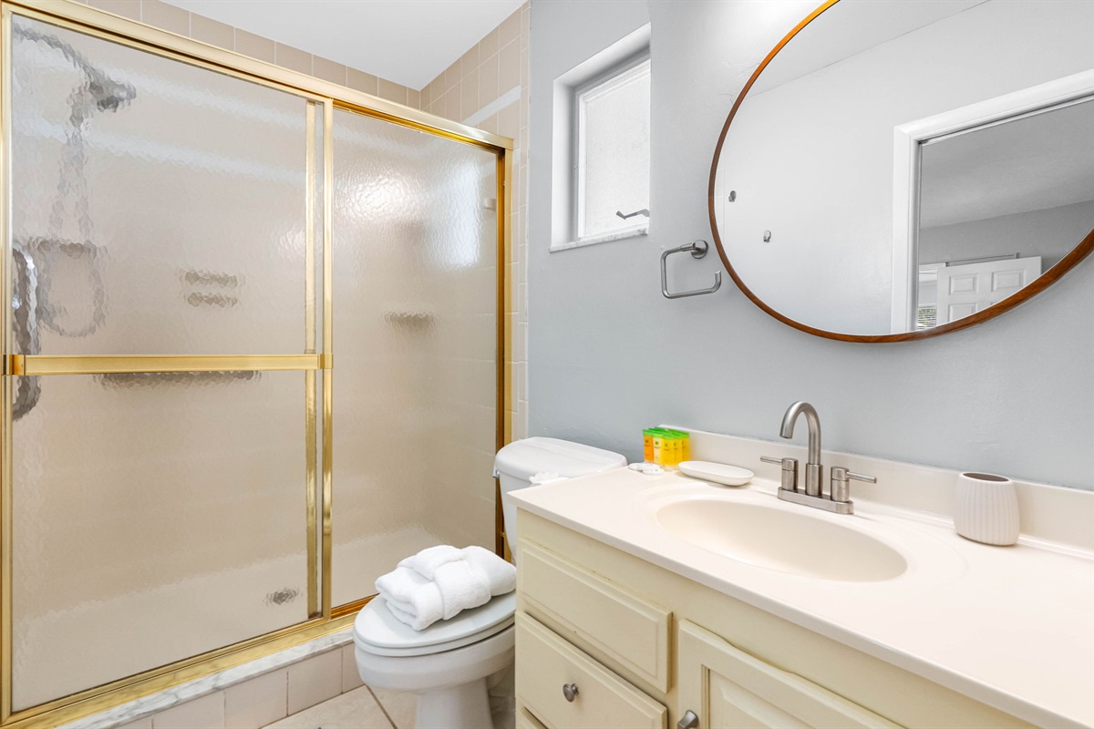 This clean and cozy bathroom features a spacious walk-in shower with gold trim, a round mirror, and natural light from the frosted window.