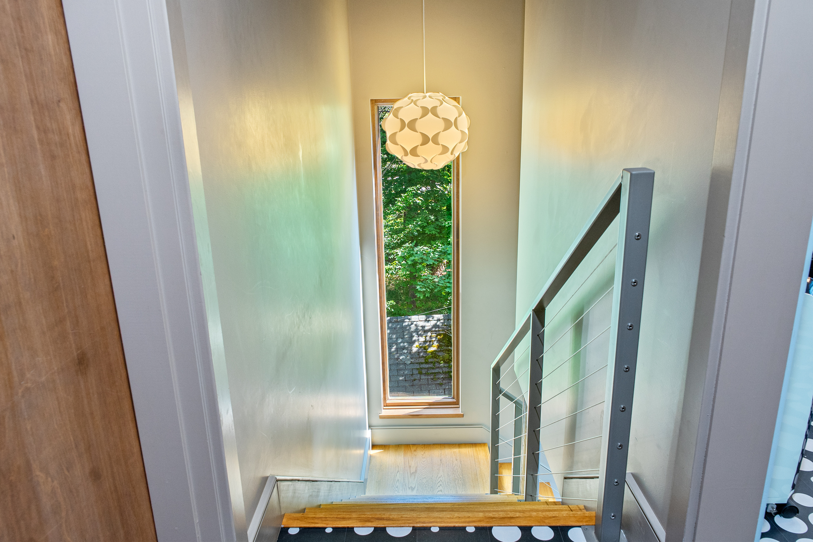 Architectural beauty in the "Sky Passage" at "The Crag." Looking down the stairwell reveals a glowing geometric pendant light, tall window framing the woods, modern cable railing, and eye-catching patterned floor details connecting the levels.