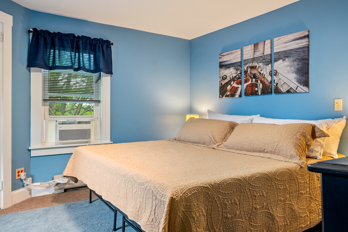 This bedside perspective highlights the premium sleeping amenities of the primary bedroom suite. Combining a spacious king memory foam mattress with dedicated in-window AC cooling and custom nautical artwork, this room prioritizes guest comfort.