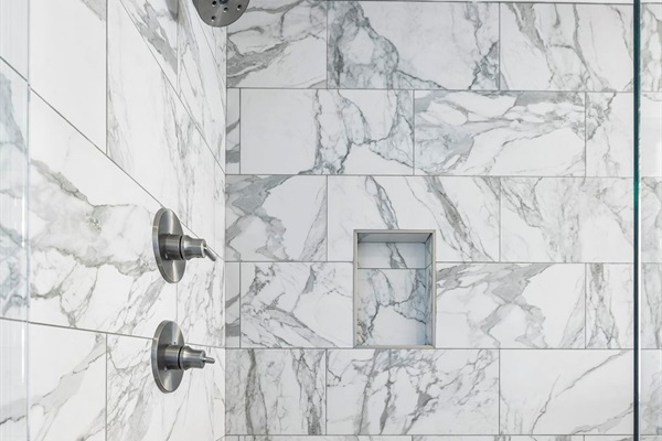 Frameless glass doors reveal stunning marble tile with soft gray veining throughout - Built in niche for convenience and multiple spray heads for a refreshing experience - Soft lighting and stone style flooring create a calming, upscale shower retreat