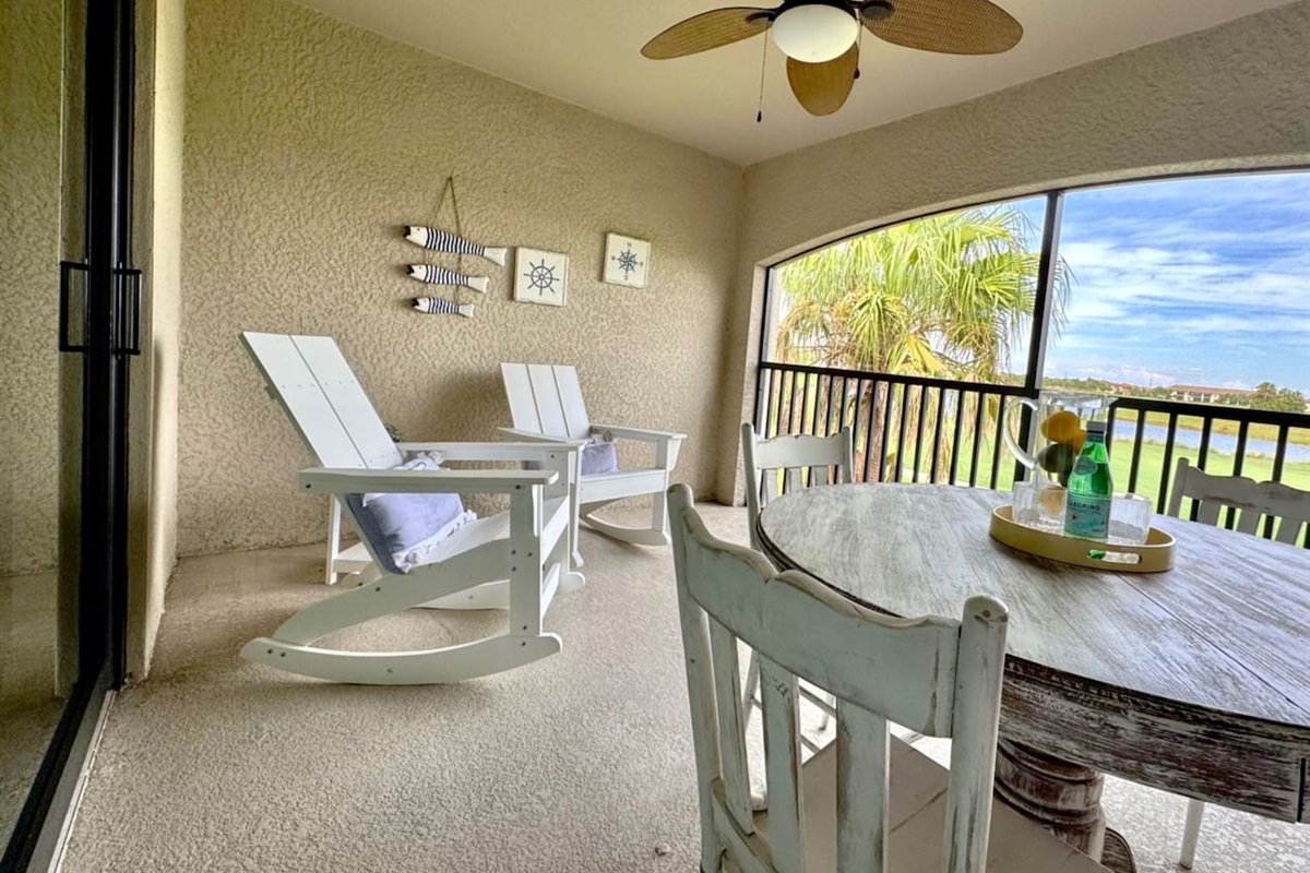 Cozy screened lanai with rocking chairs, round table with 4 chairs. The view outside shows palm trees, a golf course, and a lake.