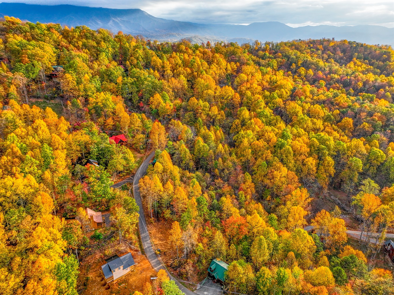 A breathtaking aerial view of a vibrant autumn forest, with golden, orange, and red hues blanketing the hills. Cozy cabins peek through the trees, and distant mountain peaks complete the stunning scene.