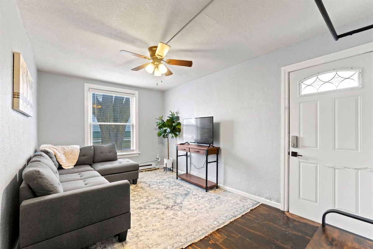 Inviting living room with a bright window, featuring a grey sectional couch with a cozy throw blanket and a TV for ultimate comfort and relaxation."