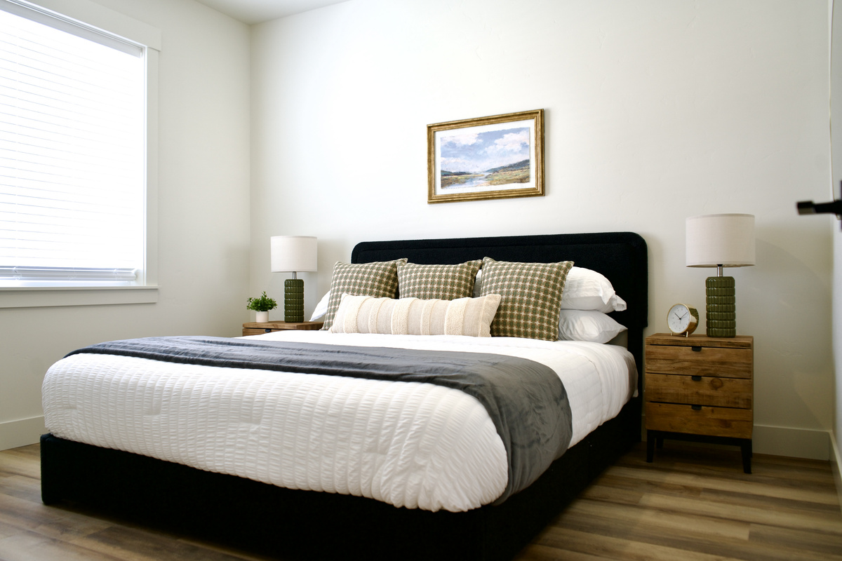 The downstairs bedroom features a comfortable queen-size bed with matching nightstands and bedside lamps on each side. With its high ceilings and spacious layout, this inviting retreat offers a bright, open atmosphere designed for comfort and relaxation.  
