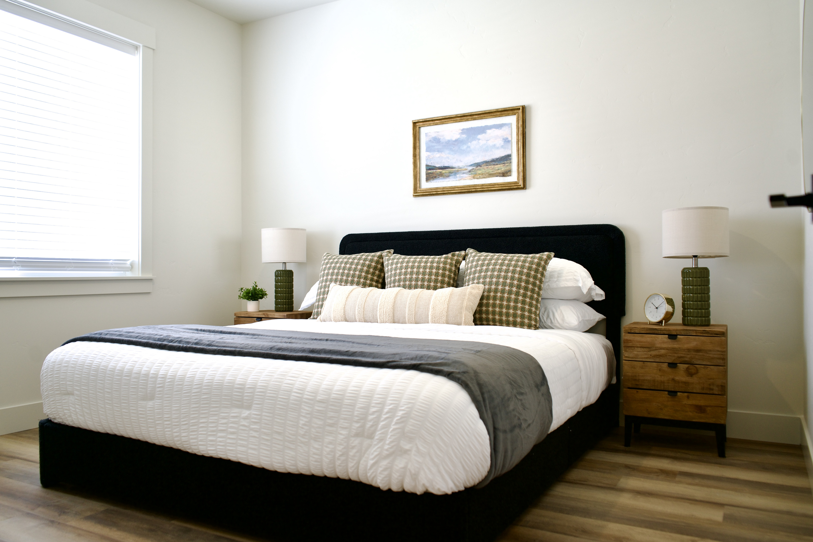 The downstairs bedroom features a comfortable queen-size bed with matching nightstands and bedside lamps on each side. With its high ceilings and spacious layout, this inviting retreat offers a bright, open atmosphere designed for comfort and relaxation.  
