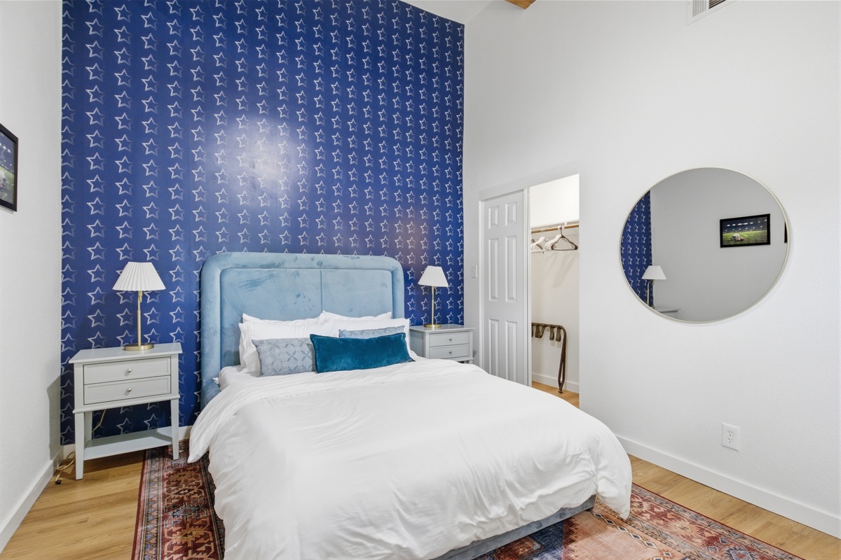 Bedroom 4 wide-angle with navy wallpaper accent wall, round mirror, and hardwood floors. Clothing storage and room-darkening shades included. Sleeps 8 total.