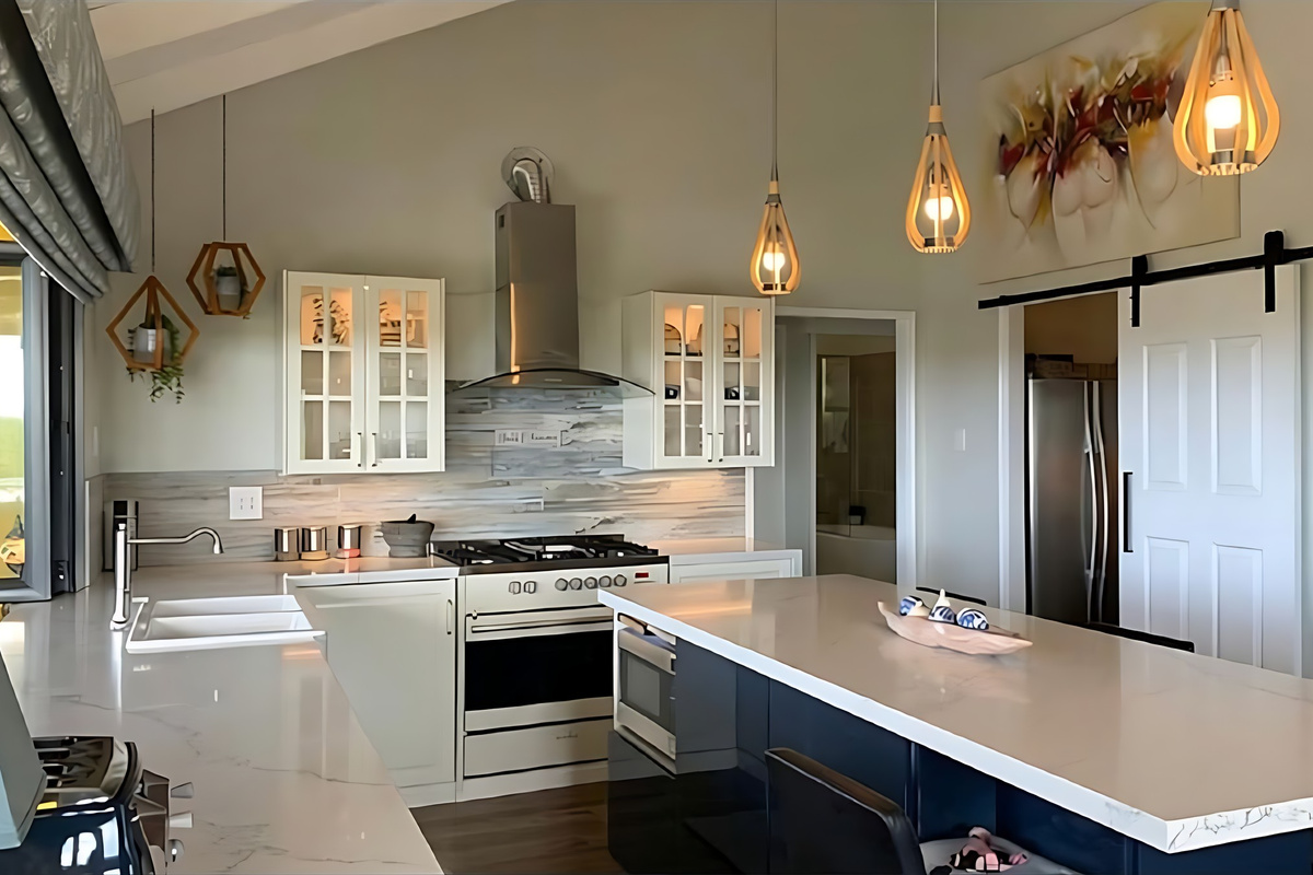 The kitchen has a raked ceiling, timber pendant lights, stone-look benchtops and a freestanding gas cooker, with a white barn door sliding through to the scullery where the fridge is kept.