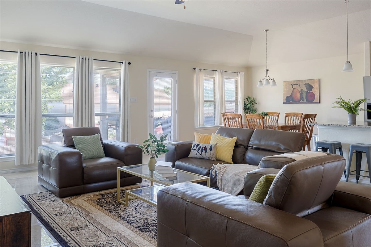 Step into a well-lit living area featuring cozy leather sofas, a chic coffee table, and expansive windows connecting to the dining area and kitchen.