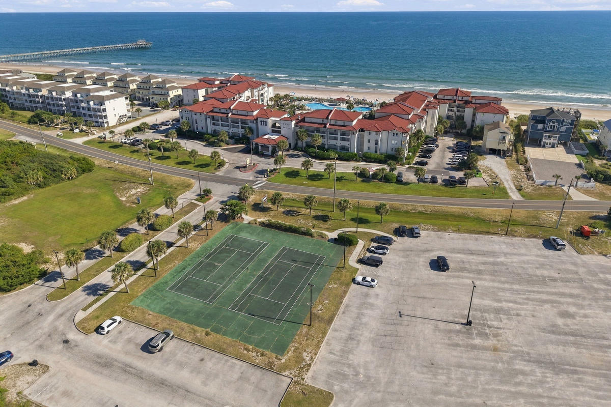 Aerial view of the resort grounds featuring outdoor tennis courts, spacious parking areas, and terracotta-roofed residential buildings bordering the oceanfront.   
