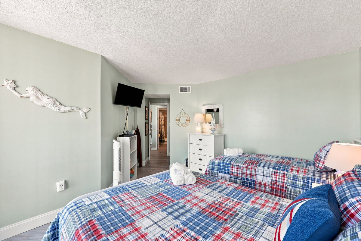 Comfortable guest bedroom featuring two beds with colorful plaid bedding, a wall-mounted flat-screen TV, and unique coastal wall art.