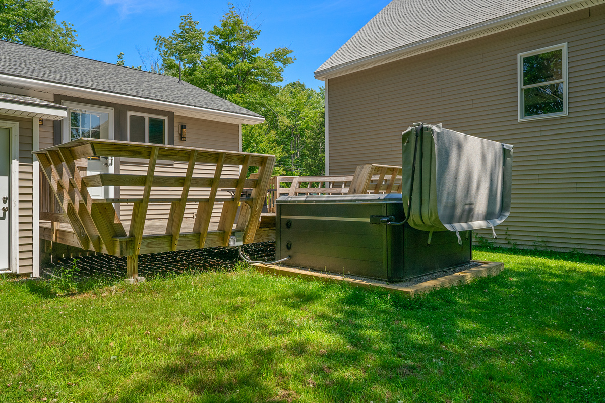 Walk from the grassy lawn onto the spacious back deck featuring built-in wooden bench seating. This side perspective highlights the private 6-person hot tub with its open cover lifter, located steps away from the kitchen and rear entry doors.