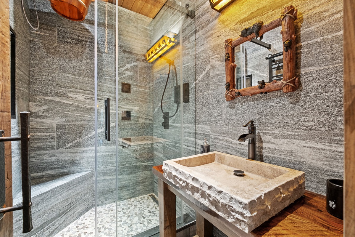 Luxury spa bathroom featuring a handcrafted rough-cut stone sink, oversized walk-in shower with pebble flooring, premium rainfall fixtures, and a traditional cold-water dump bucket for the ful