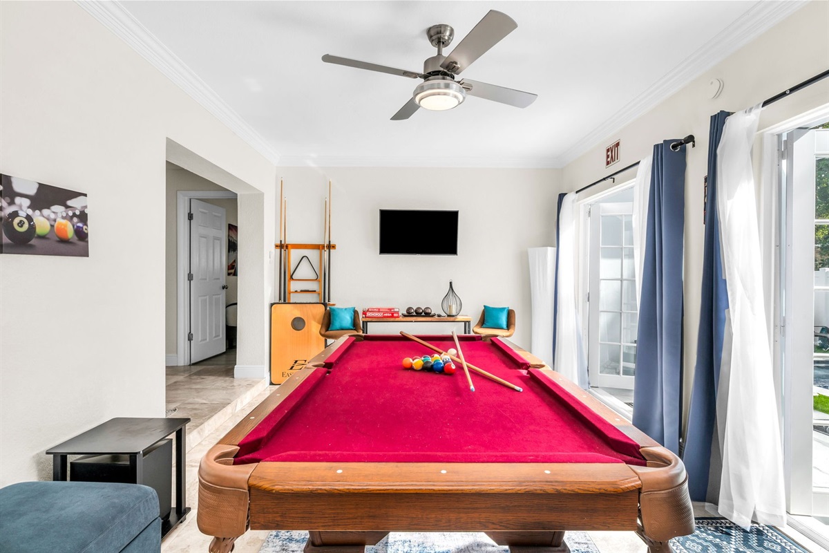 Bright Florida sunroom with a full-size pool table, seating area, beverage cooler, and a streaming TV. French doors open directly to the pool deck for seamless indoor–outdoor entertainment.